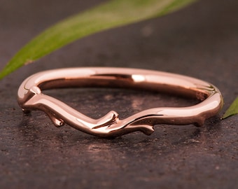 Puede incluir: Un anillo de oro rosa con un diseño inspirado en la naturaleza. El anillo presenta una banda curva con un motivo en forma de rama, que incluye pequeños detalles texturizados. El anillo se encuentra sobre un fondo oscuro con hojas verdes.