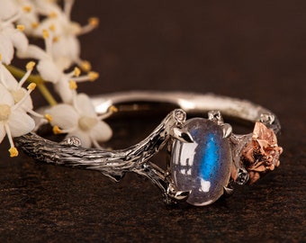 May include: A silver ring with a blue labradorite gemstone set in a branch-like design. The ring also features a rose-shaped detail on the side.