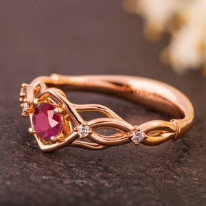 May include: A rose gold engagement ring with a ruby center stone and diamond accents. The ring features a delicate, intertwined design.