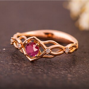 May include: A rose gold ring with a ruby gemstone surrounded by smaller diamonds. The ring features a delicate, interwoven design.