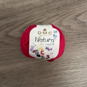 May include: A ball of red Natura Just Cotton yarn with a white label featuring the DMC logo and floral illustrations. The label also includes the text "Natura Just Cotton". This yarn is suitable for various crafting projects.
