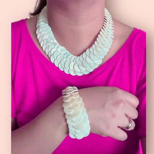 May include: A white shell necklace and bracelet set. The necklace is made of many small, round shells that are layered on top of each other. The bracelet is made of the same type of shells, but it is smaller than the necklace.