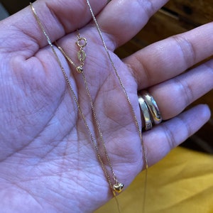 May include: Two gold necklaces, one with a small heart pendant and the other with a small circle pendant. Both necklaces are on a hand.