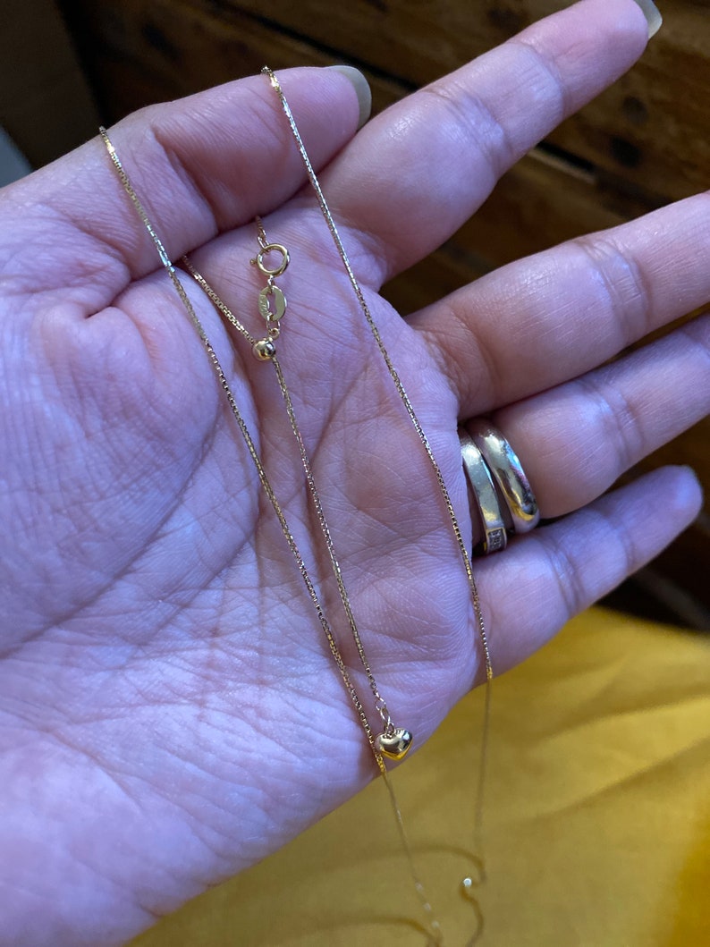 May include: Two gold necklaces, one with a small heart pendant and the other with a small circle pendant. Both necklaces are on a hand.