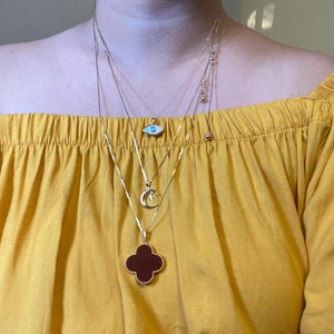 May include: A woman is wearing four gold necklaces with different pendants. The pendants include a blue evil eye, a crescent moon, and a four-leaf clover with a red stone.