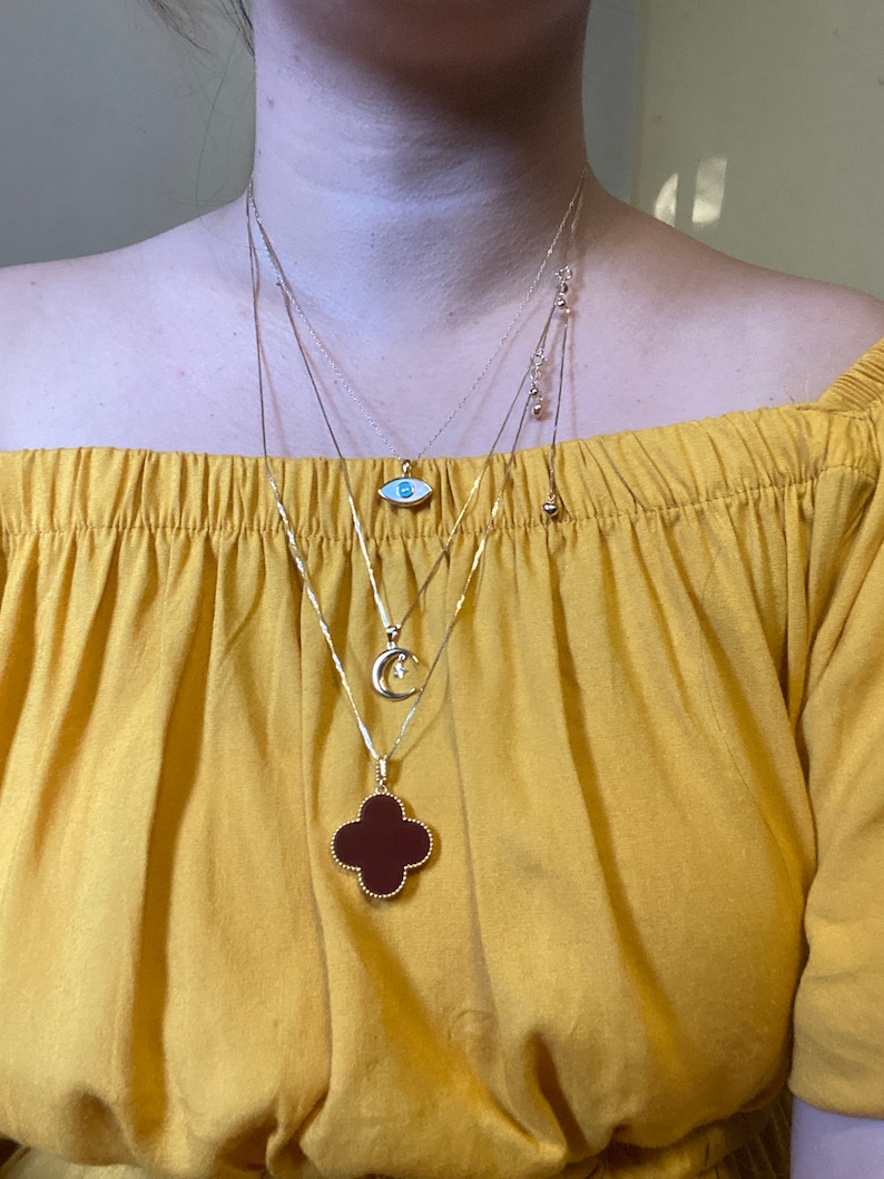 May include: A woman is wearing four gold necklaces with different pendants. The pendants include a blue evil eye, a crescent moon, and a four-leaf clover with a red stone.