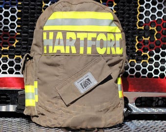 Könnte beinhalten: Ein beigefarbener Rucksack aus strapazierfähigem Stoff mit horizontalen gelben Reflexstreifen und dem Wort HARTFORD in großen gelben Buchstaben. Auf der Vordertasche befindet sich ein rechteckiger Aufnäher mit dem Text MORNING PRIDE.