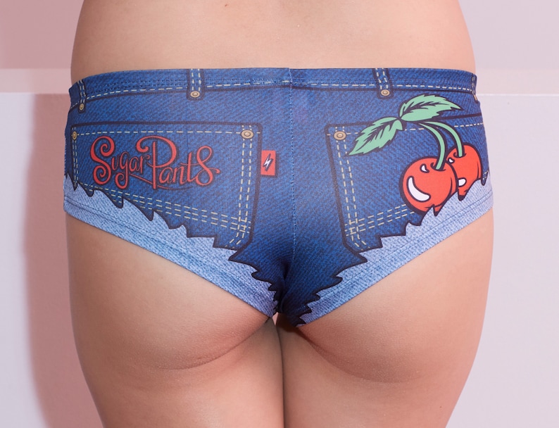 Pu&ograve; includere: Intimo in denim blu con un aspetto consumato e una tasca con il testo "Sugar Pants" e una grafica di una ciliegia.