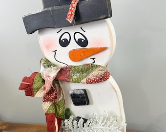 May include: Wooden snowman decoration with a black top hat, carrot nose, and a patterned scarf in red, green, and white. The snowman has a painted face and is decorated with a bow and a pinecone.