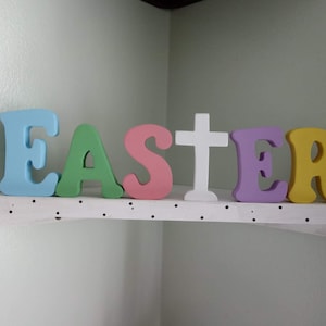May include: A white shelf with black polka dots displays the word "EASTER" in colorful wooden letters. The letters are painted in shades of blue, green, pink, purple, and yellow. A white wooden cross is between the "S" and "T".