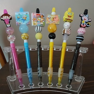May include: A collection of decorative pens in a clear acrylic holder. Each pen features a different cartoon character charm, including Dora the Explorer, SpongeBob SquarePants, and Minions. The pens have colorful barrels and decorative beads.