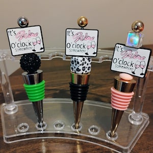 May include: Three decorative wine bottle stoppers with silver, black, and pink accents. Each stopper has a unique design, including a black beaded ball, a black and white leopard print, and a pink striped design. Each stopper has a small sign that says "It's Wine O'clock Somewhere."