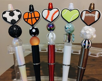 Puede incluir: Una colección de bolígrafos decorativos con adornos temáticos deportivos. Cada bolígrafo presenta un deporte diferente: fútbol, baloncesto, béisbol, tenis y fútbol americano. Los bolígrafos tienen varios colores y diseños, incluyendo negro, blanco, rojo y marrón.