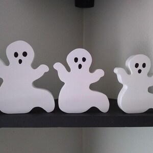 May include: Three white wooden ghost figurines with black eyes and mouths. The ghosts are arranged in a row on a black shelf.