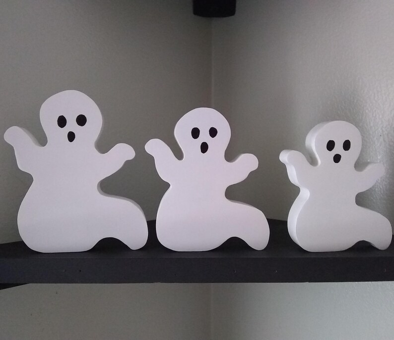 May include: Three white wooden ghost figurines with black eyes and mouths. The ghosts are arranged in a row on a black shelf.