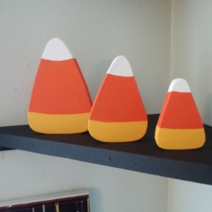May include: Three wooden candy corn decorations in different sizes. The largest is orange, yellow, and white. The medium is orange, yellow, and white. The smallest is orange and yellow.