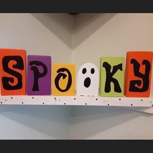 May include: Wooden blocks spelling out the word "Spooky" in black letters. The blocks are painted in various colors: orange, purple, yellow, white, green, and orange. A ghost is depicted on the white block.
