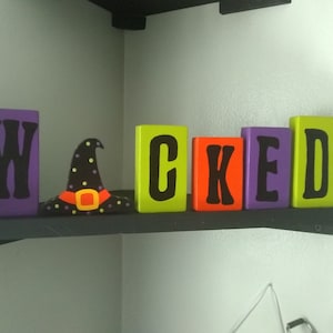May include: Wooden blocks spelling out the word "WICKED" in black letters on a purple, green, orange, and yellow background. A black witch's hat with green and yellow accents sits in front of the blocks.