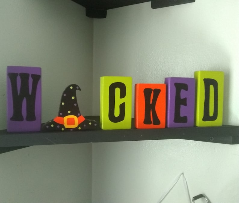 May include: Wooden blocks spelling out the word "WICKED" in black letters on a purple, green, orange, and yellow background. A black witch's hat with green and yellow accents sits in front of the blocks.