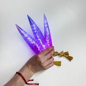 May include: A hand holds a decorative, illuminated object with three purple, leaf-like shapes. The tips are blue, and the object has small lights inside. A gold tassel hangs from the base, and a red bracelet is on the wrist.