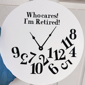 May include: White round clock with black hands and black numbers. The numbers are scattered around the clock face. The clock face says "Who cares! I'm Retired!"