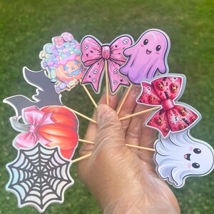 May include: A collection of decorative cupcake toppers featuring Halloween-themed illustrations including a pumpkin with candy, a pink bow with skull patterns, a smiling pink ghost, a red pumpkin with a pink bow, a spider web, and a white ghost.