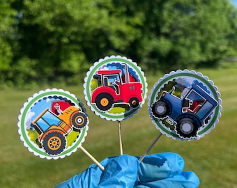 May include: Three round paper cupcake toppers with a green border and scalloped edges. Each topper features a cartoon tractor with a different color: red, yellow, and blue. The tractors are on a green background with a farm scene.