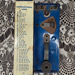 May include: Vintage blue and cream International Code telegraph key. The cream-colored panel displays the Morse code alphabet and numbers. The blue metal base has a lever and a circular component. The item shows signs of age.