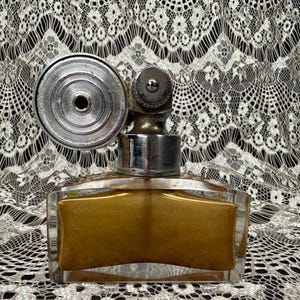 May include: A vintage perfume bottle with a rectangular, clear glass body and a silver-toned atomizer. The bottle contains a golden-colored liquid. The atomizer has a circular design and a small, rounded top. The background is a black and white lace.