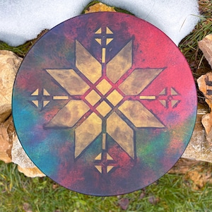 May include: A round, decorative object with a starburst design. The object features a gold star in the center, surrounded by a red, blue, and green background. The object is placed on a bed of grass and rocks.