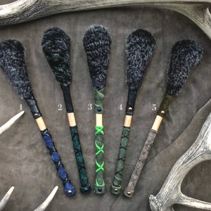 Op de afbeelding: Vijf grijze en zwarte bontpompoenen bevestigd aan houten stokken met leren omwikkelingen. De leren omwikkelingen zijn in verschillende kleuren en patronen, waaronder groen, blauw en zwart.