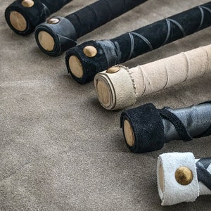 May include: Close-up of several handcrafted wooden batons with leather-wrapped handles. The batons feature black, white, and tan leather, with decorative stitching and brass accents. The batons are arranged on a textured grey surface.