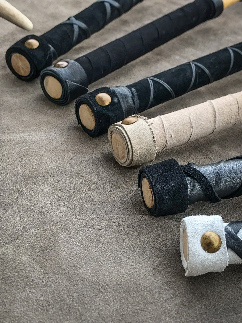 May include: Close-up of several handcrafted wooden batons with leather-wrapped handles. The batons feature black, white, and tan leather, with decorative stitching and brass accents. The batons are arranged on a textured grey surface.