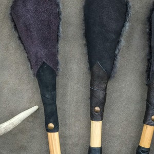 May include: Three handmade leather and fur paddles are displayed. Each paddle features a wooden handle wrapped in black leather, and a fur-lined head. The paddle heads are dark grey and black. A white antler is visible.