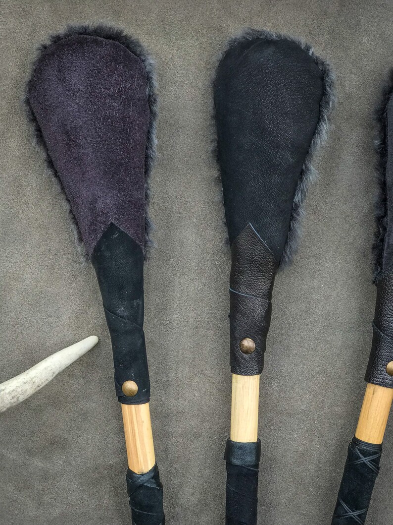 May include: Three handmade leather and fur paddles are displayed. Each paddle features a wooden handle wrapped in black leather, and a fur-lined head. The paddle heads are dark grey and black. A white antler is visible.