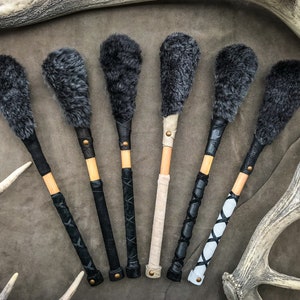 May include: Six drum beaters with gray fur heads and wooden handles. The handles are wrapped in black, beige, and white leather with decorative stitching and brass rivets. The beaters are displayed on a brown surface.