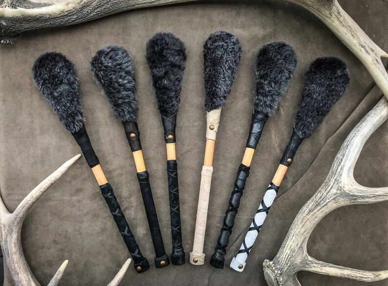 May include: Six drum beaters with gray fur heads and wooden handles. The handles are wrapped in black, beige, and white leather with decorative stitching and brass rivets. The beaters are displayed on a brown surface.