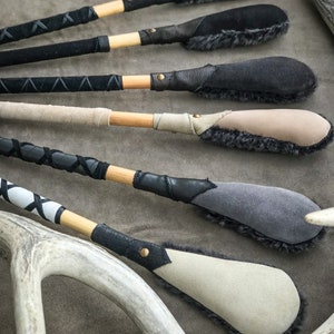 May include: A selection of handcrafted leather and suede paddles with wooden handles. The paddles feature various colours, including black, grey, and beige, with decorative stitching and fur trim. Antlers are visible in the background.