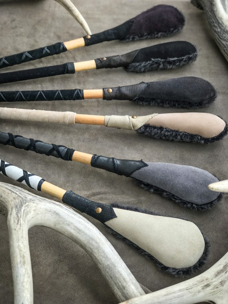 May include: A selection of handcrafted leather and suede paddles with wooden handles. The paddles feature various colours, including black, grey, and beige, with decorative stitching and fur trim. Antlers are visible in the background.