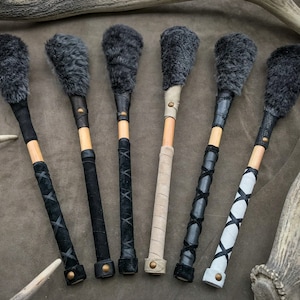 May include: Six percussion mallets with dark grey fur heads, wooden shafts, and leather-wrapped handles. The handles feature various leather designs in black, tan, and white, secured with brass rivets. The mallets are arranged on a brown surface.