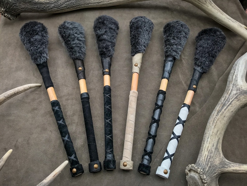 May include: Six percussion mallets with dark grey fur heads, wooden shafts, and leather-wrapped handles. The handles feature various leather designs in black, tan, and white, secured with brass rivets. The mallets are arranged on a brown surface.