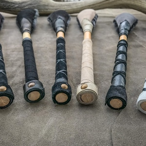 May include: Six handcrafted wooden clubs with leather-wrapped handles and suede heads. The handles are wrapped in black, grey, and tan leather, with decorative stitching. The heads are a mix of black, grey, and tan suede. Each club has a wooden base.