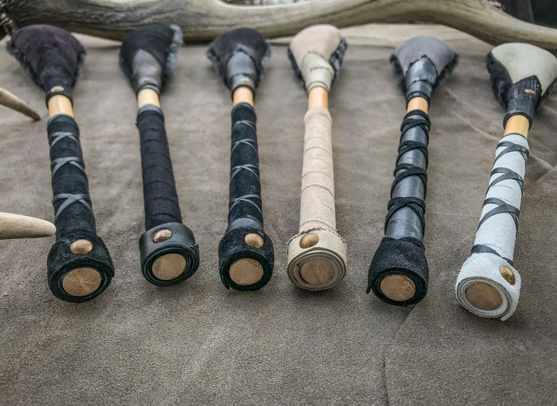 May include: Six handcrafted wooden clubs with leather-wrapped handles and suede heads. The handles are wrapped in black, grey, and tan leather, with decorative stitching. The heads are a mix of black, grey, and tan suede. Each club has a wooden base.