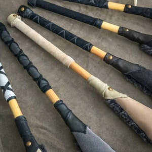 May include: A selection of handcrafted wooden paddles. The handles are wrapped in black, tan, and white leather with decorative stitching. The paddle heads are in shades of grey and tan, with suede or fur detailing.