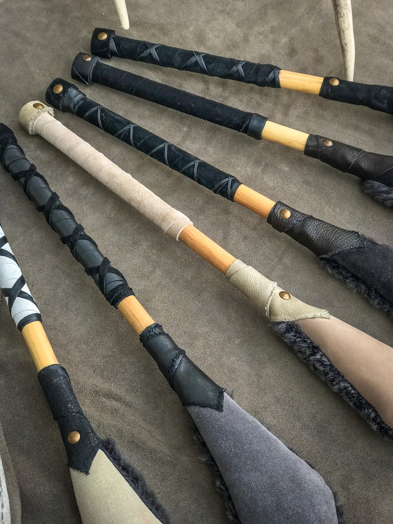 May include: A selection of handcrafted wooden paddles. The handles are wrapped in black, tan, and white leather with decorative stitching. The paddle heads are in shades of grey and tan, with suede or fur detailing.