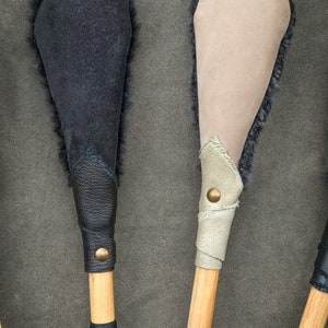 May include: Close-up of two handcrafted wooden mallets. One mallet features a black suede head with dark fur trim, and the other has a tan suede head with fur trim. Both have leather-wrapped handles with brass button accents.