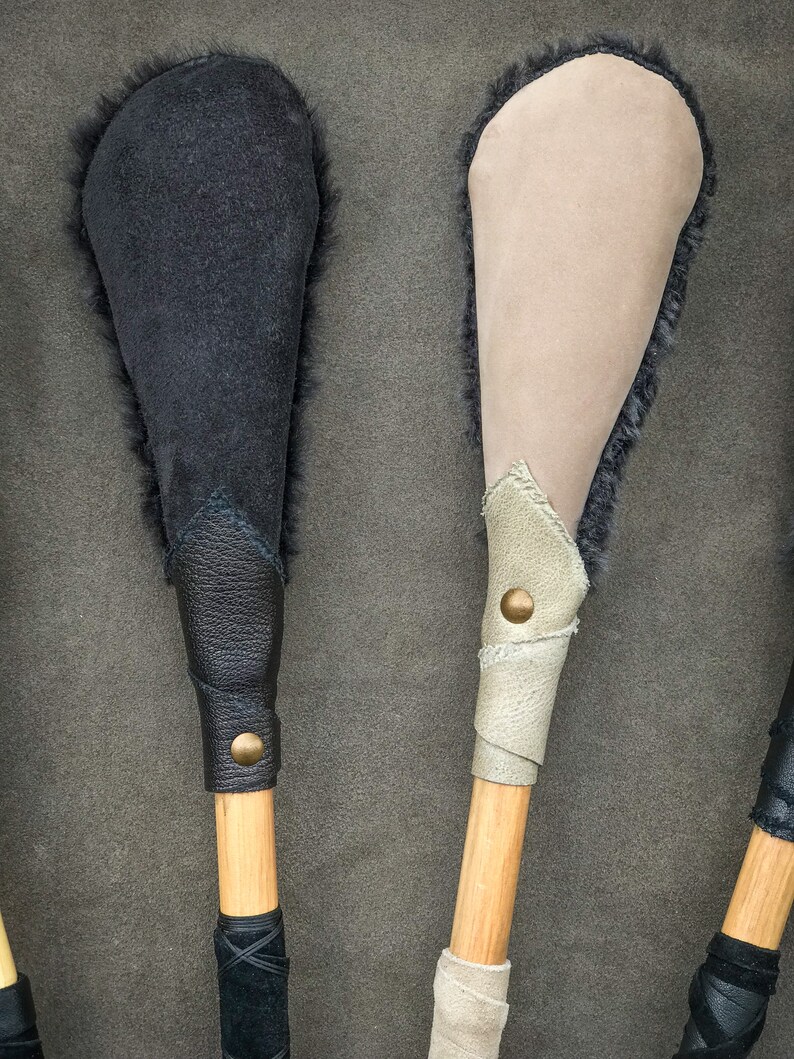 May include: Close-up of two handcrafted wooden mallets. One mallet features a black suede head with dark fur trim, and the other has a tan suede head with fur trim. Both have leather-wrapped handles with brass button accents.