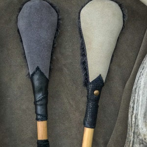 May include: Two handcrafted drum beaters. One has a grey suede head with black fur trim, a black leather handle, and a wooden shaft. The other has a cream suede head with black fur trim, a black leather handle, and a wooden shaft.
