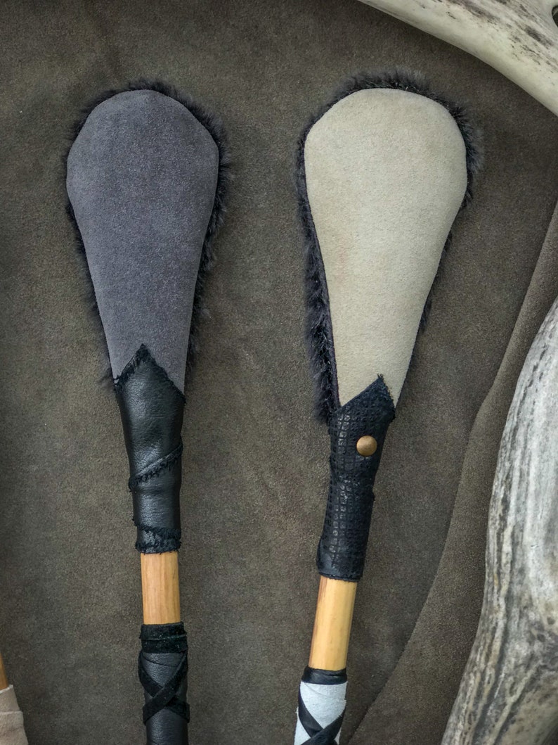 May include: Two handcrafted drum beaters. One has a grey suede head with black fur trim, a black leather handle, and a wooden shaft. The other has a cream suede head with black fur trim, a black leather handle, and a wooden shaft.