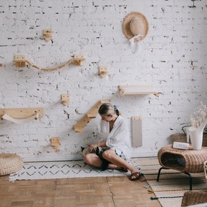May include: A white brick wall with a cat climbing wall made of wood. The wall features shelves, ramps, and a rope bridge. A woman is sitting on the floor with a cat in her lap.
